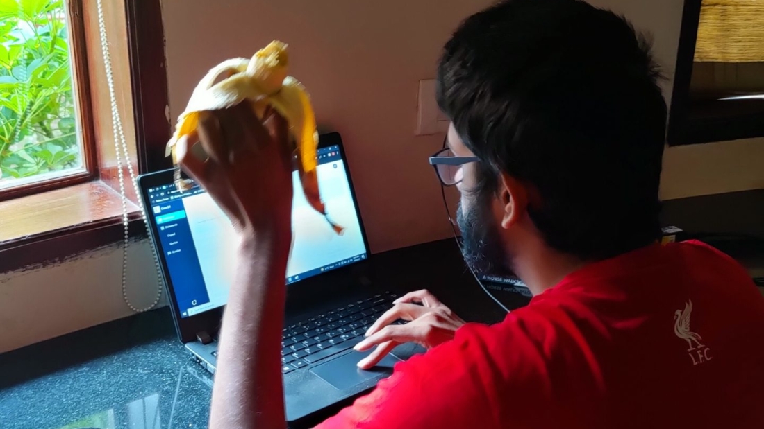 A man sitting and working on a laptop, and eating a banana